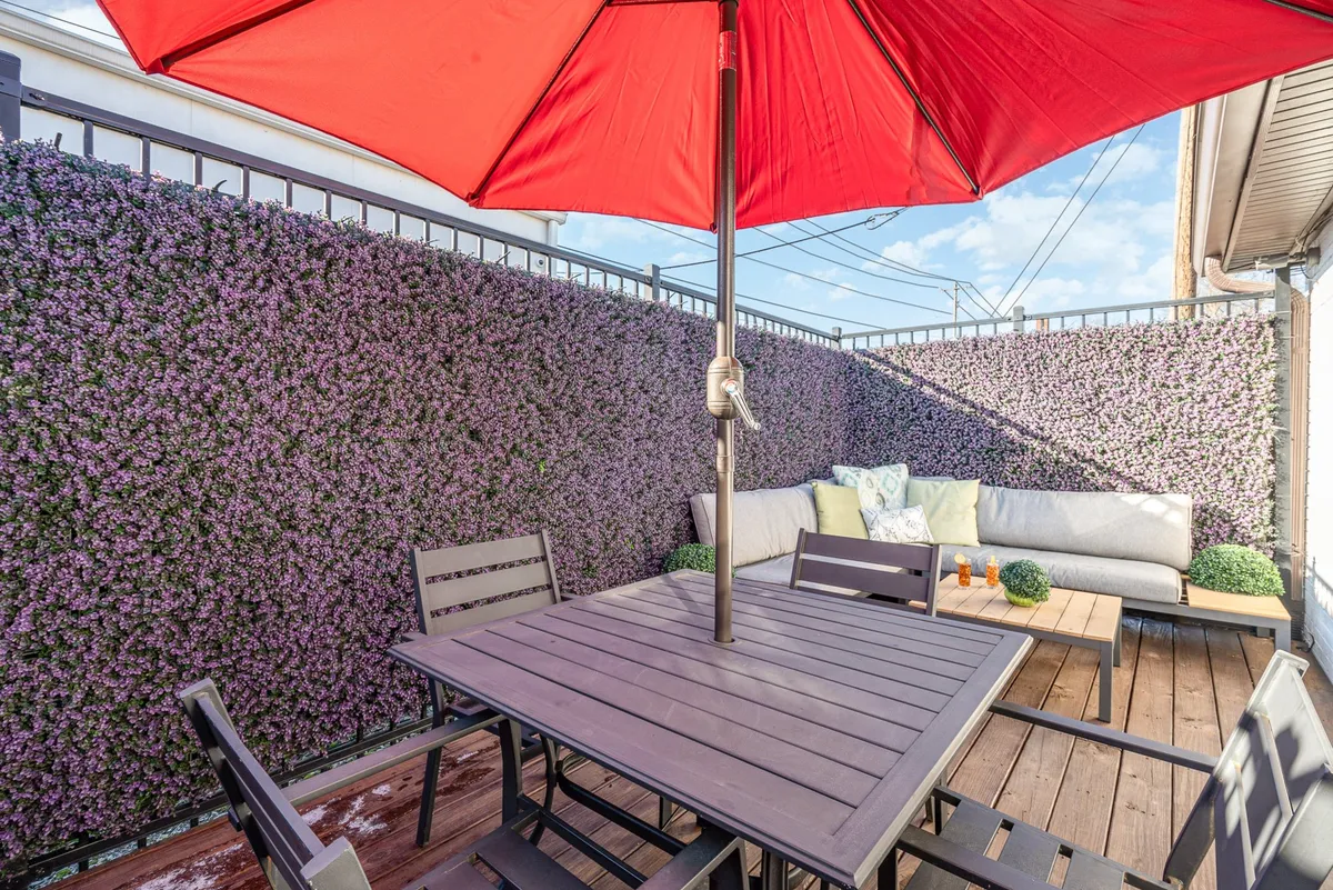 This scene showcases an exterior patio with a red umbrella, outdoor seating, a purple accent wall, and a coffee table. The arrangement is perfect for group gatherings, sunny brunches, or evening conversations under the stars.