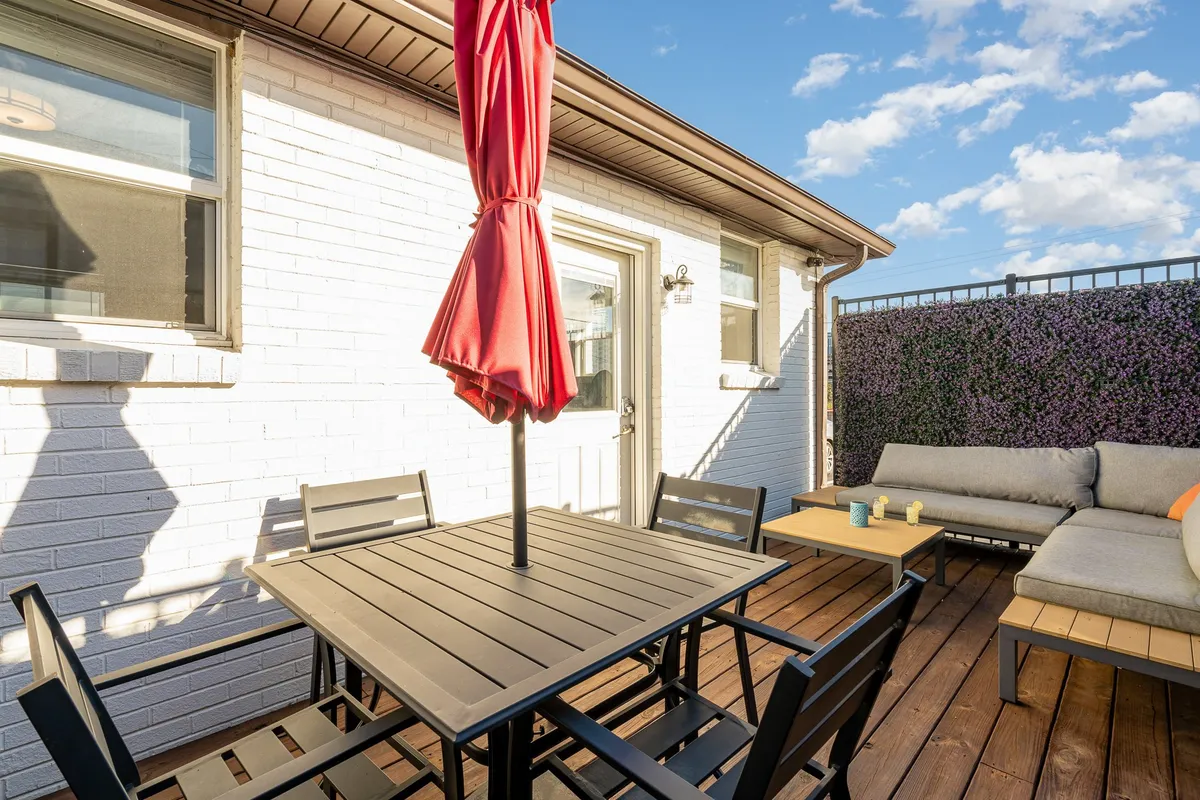 An outdoor patio scene featuring a red umbrella, a spacious dining table, and plush seating that invites relaxation and socializing. The setup suggests a comfortable place to unwind or host informal meetings.