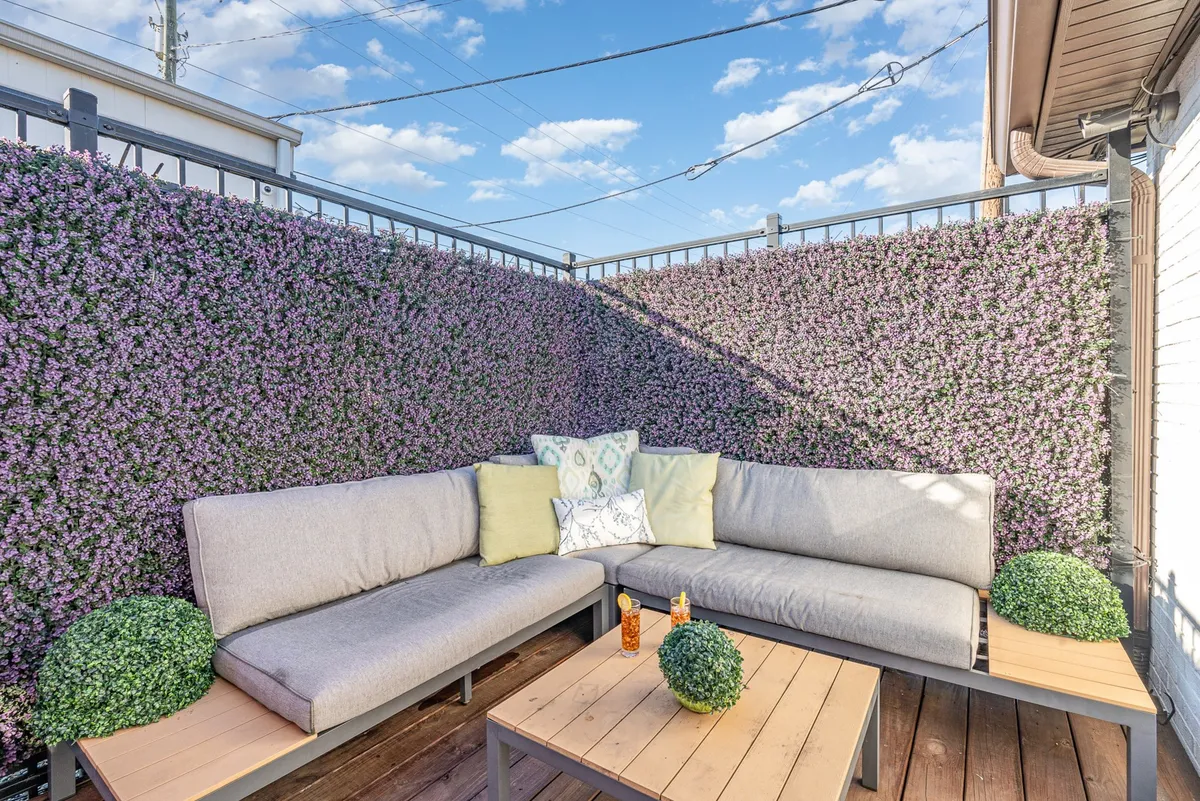 This image depicts a calm and welcoming patio with sofa seating, decorative cushions, a table, greenery, and a beautiful flowering wall. The space feels serene and is suited for quiet coffee mornings, reading, or intimate gatherings.
