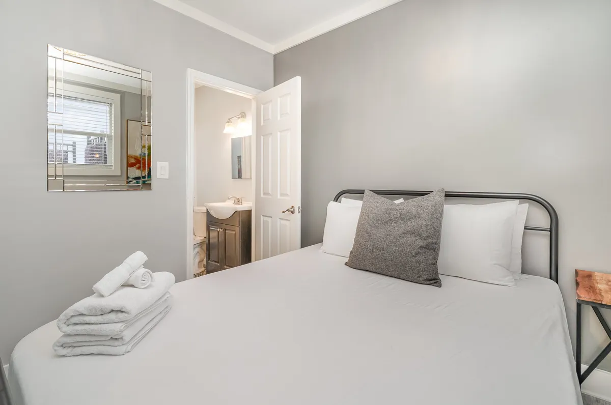 A cozy bedroom with neatly arranged pillows and towels on the bed, a mirror reflecting soft light and space, and an open doorway that clearly leads to a bright bathroom, suggesting the Jack-n-Jill layout.