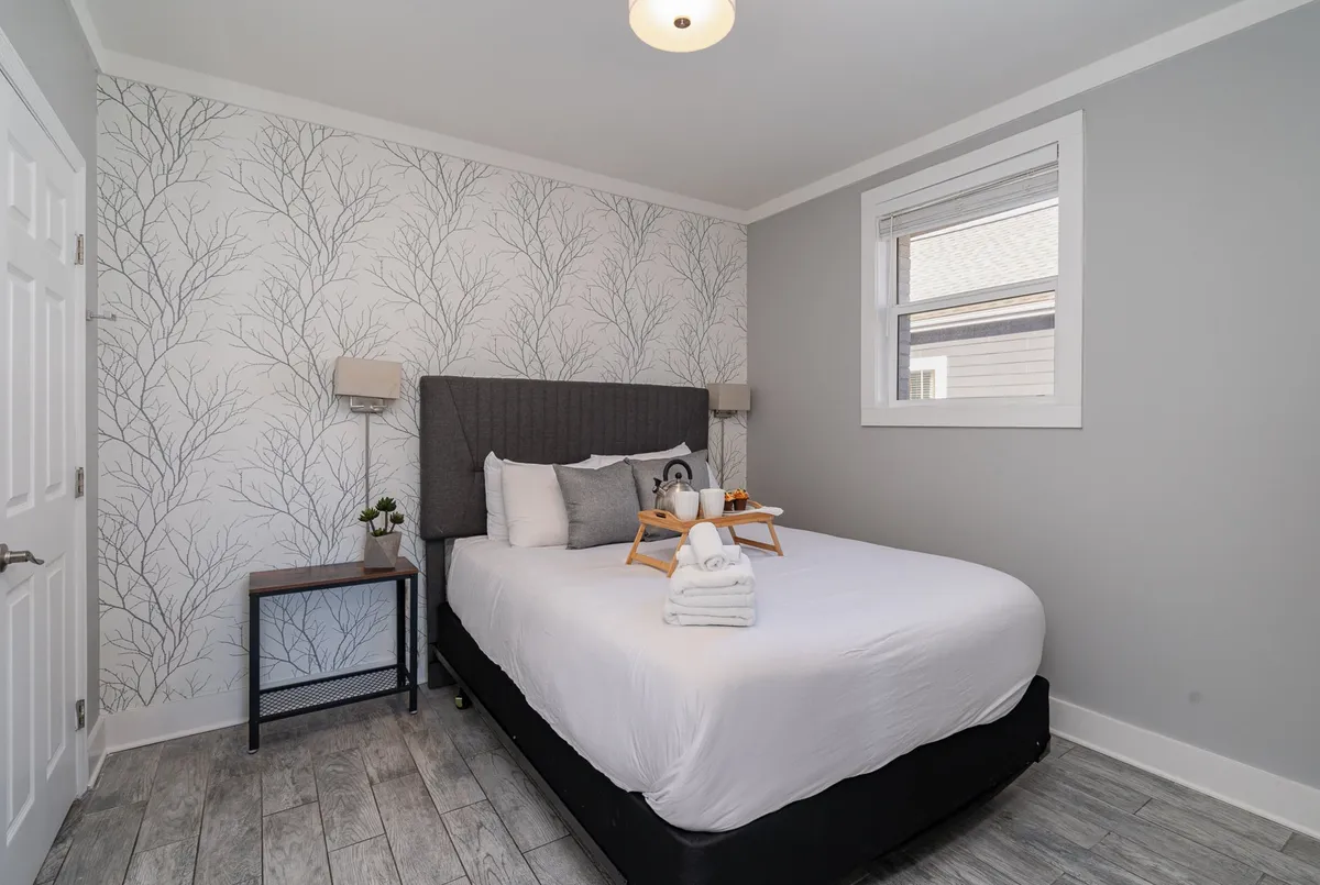 A tranquil bedroom featuring a queen bed adorned with decorative pillows, a wall art depicting tree branches, a bedside table with a lamp and small plant, and a tray with beverages, all bathed in gentle morning light.